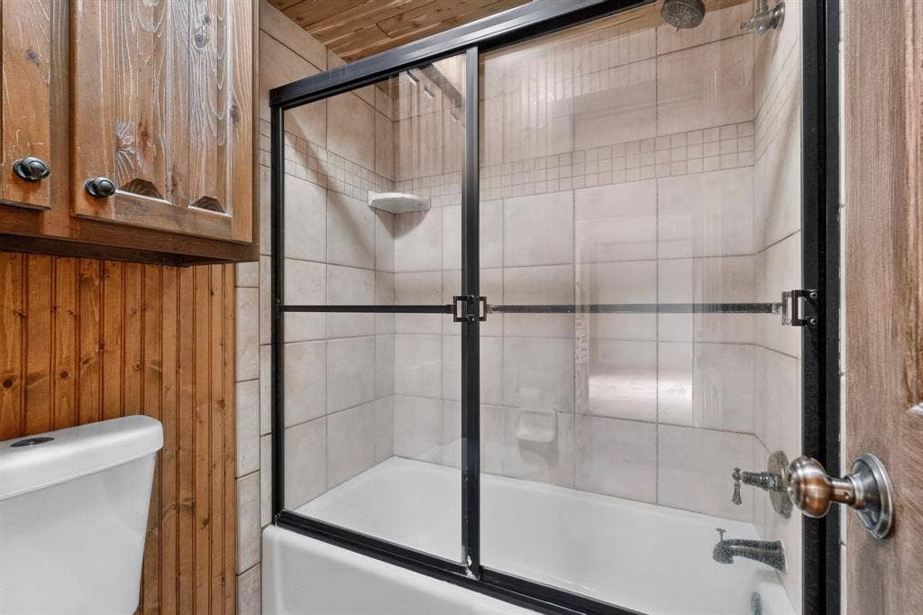 Bathtub with sliding glass shower door, light-toned tile surround, and built-in corner shelf