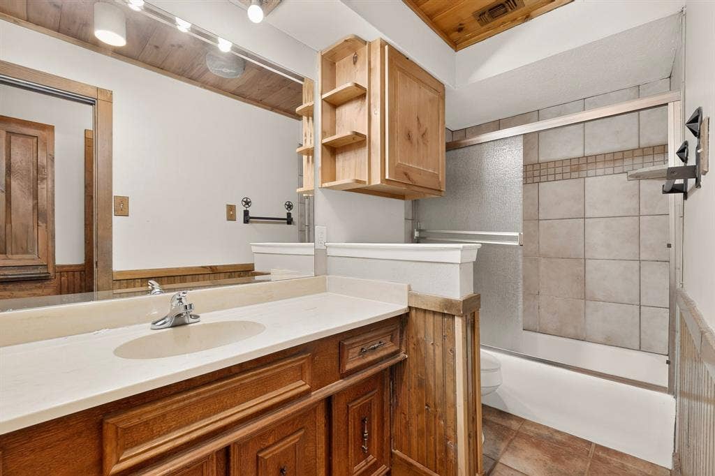 Bathroom featuring a wood-finish vanity with a light-colored countertop, an integrated sink, a large wall mirror, and a tiled shower/tub combination with sliding glass doors