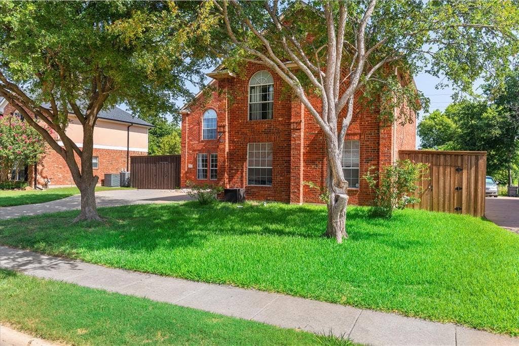 1013 Larkspur Drive, Allen, TX 75002 Red brick exterior featuring arched and rectangular window designs, a paved driveway, mature trees, and a wooden privacy fence