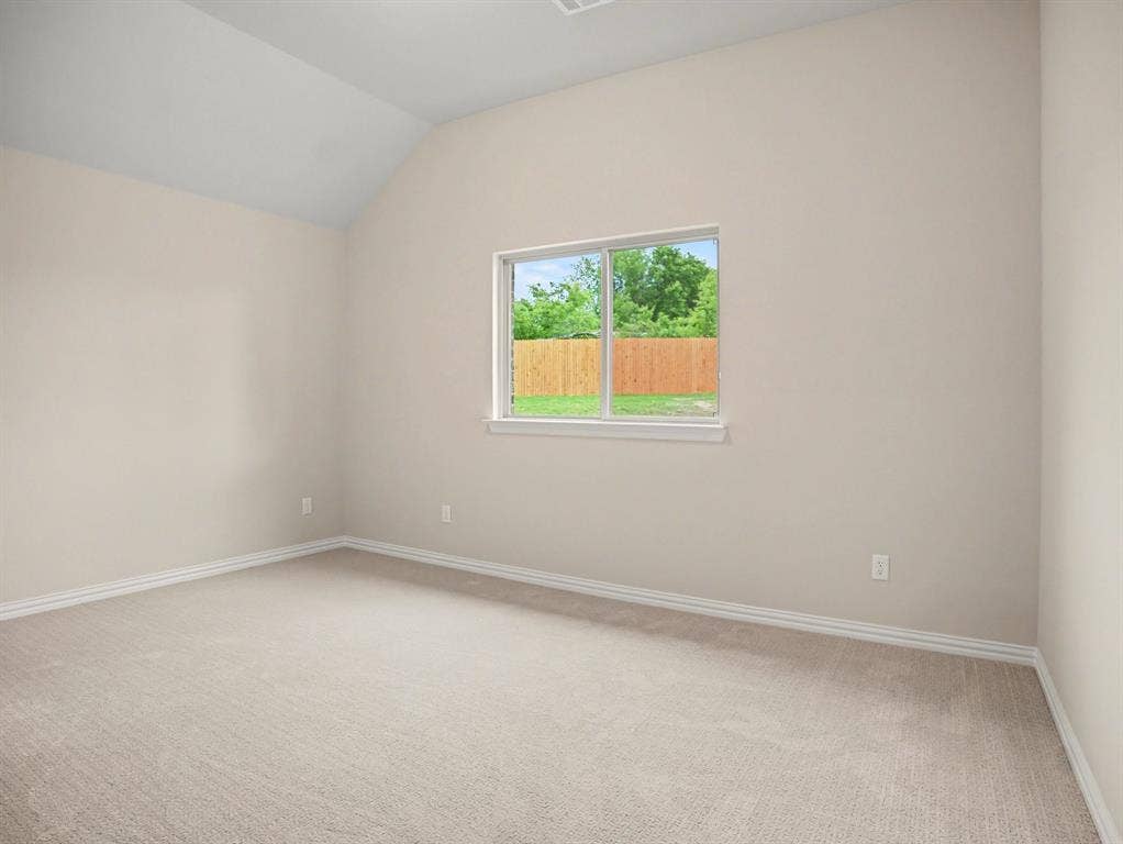Vaulted ceiling bedroom featuring plush neutral carpeting, white baseboards, and a large window with a wood fence backdrop