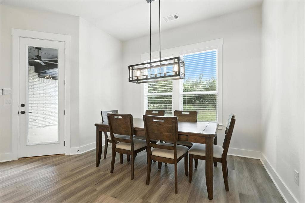 3141 Claire Drive, Midlothian, TX 76065 Dining area featuring wood-finish flooring, a contemporary rectangular chandelier, a large window with blinds, and a glass-paneled door leading to an outdoor patio with white brickwork