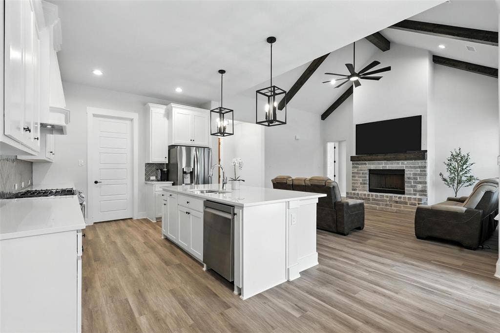 3141 Claire Drive, Midlothian, TX 76065 Open-concept living space featuring a kitchen island with sink, stainless steel appliances, wood-finish flooring, a vaulted ceiling with exposed beams, and a stone fireplace