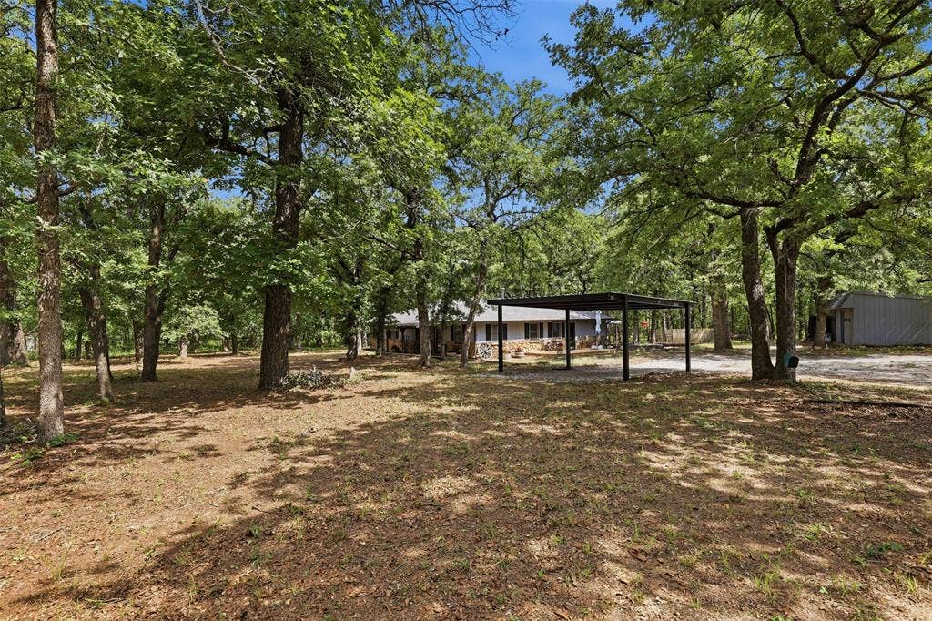 777 Wildwood Trail, Weatherford, TX 76085 Wooded lot with mature trees, a single-story home, a detached metal carport, a separate outbuilding, and an unpaved driveway