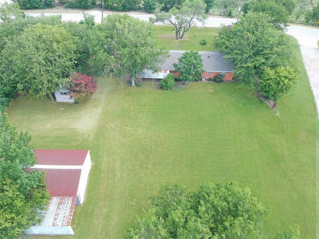 8769 S Fm 148, Scurry, TX 75158 Expansive green lawn bordering a paved road, featuring a single-story residence with a dark roof and brick exterior, mature trees, and a separate structure with a red metal roof