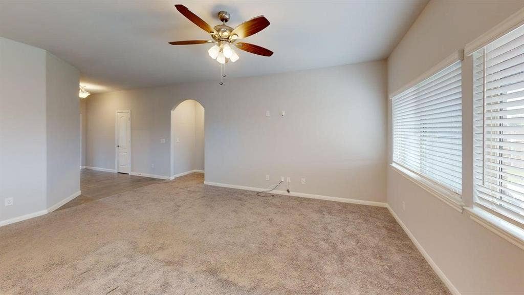 Expansive carpeted room featuring a ceiling fan with integrated lighting, two windows with blinds, and an arched doorway