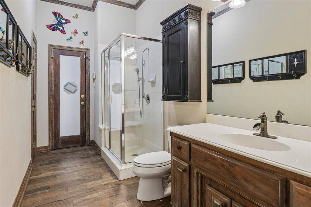 2600 Saratoga, McKinney, TX 75070 Wood-finish flooring, glass-enclosed shower, dark wood vanity with white countertop, wall-mounted dark wood cabinet, and full-width mirror