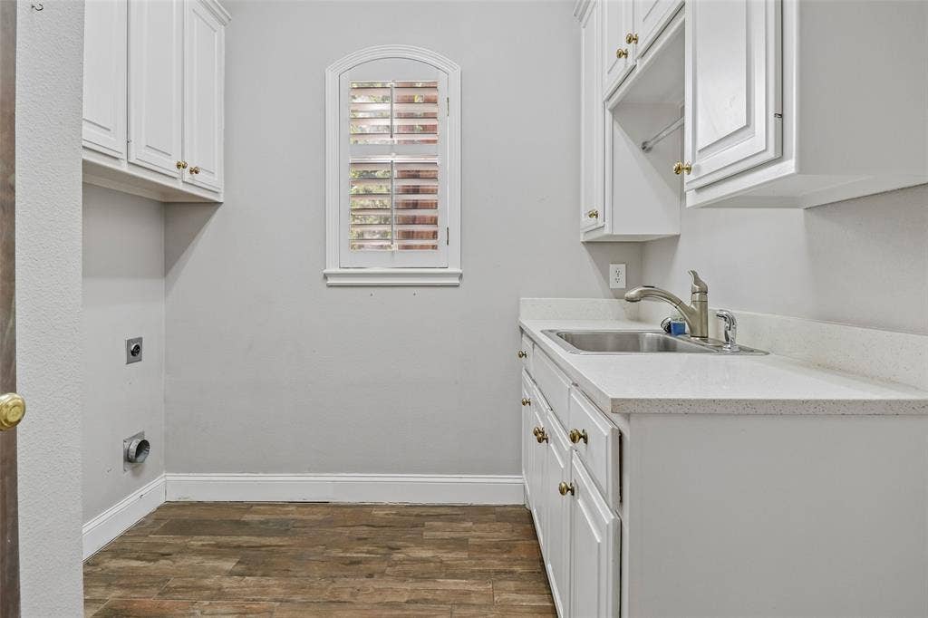 2600 Saratoga, McKinney, TX 75070 White cabinetry with brass hardware, undermount stainless steel sink, light-toned countertop, wood-finish flooring, and an arched window with shutters