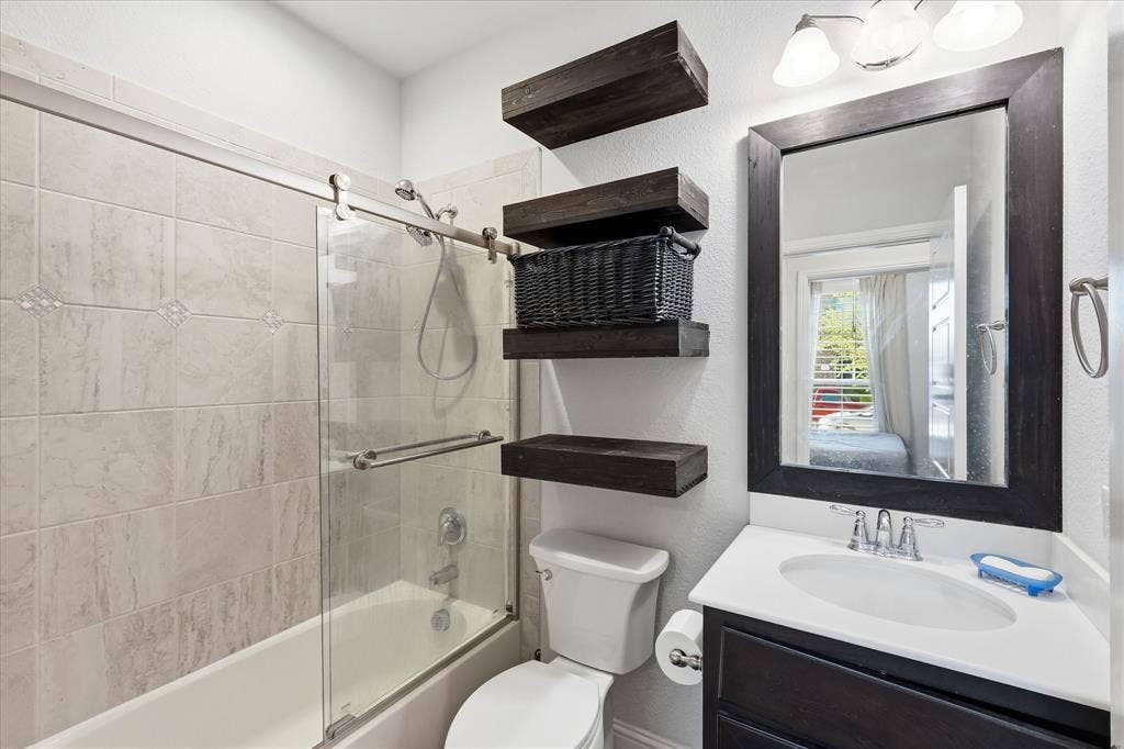 904 10th Street, Argyle, TX 76226 Bathroom featuring a tub/shower combination with sliding glass doors and tiled surround, a dark wood-finish vanity with an integrated sink and chrome faucet, a rectangular mirror with a dark wood-finish frame, and floating wood-finish shelves