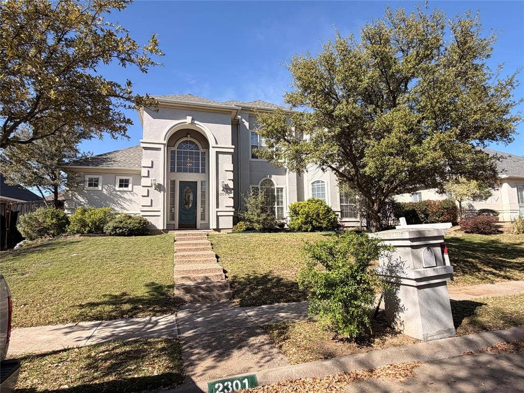 2301 Clearspring Drive S, Irving, TX 75063 Stucco exterior featuring an arched entryway with a decorative glass door, a stone-finish walkway, mature trees, and a manicured lawn