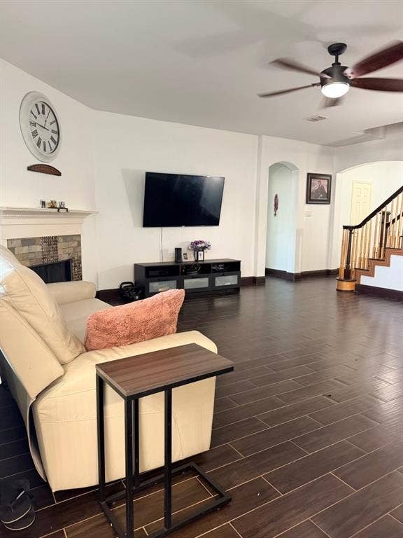 Spacious living area featuring wood-look tile flooring, a fireplace with a stone surround and white mantel, a ceiling fan with integrated lighting, and an arched entryway
