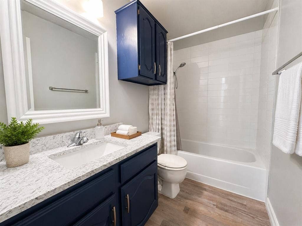 601 Ascot Drive, Euless, TX 76040 Bathroom featuring a vanity with a white countertop and dark blue cabinetry, a built-in tub with white subway tile surround, a wall-mounted mirror, and wood-finish flooring