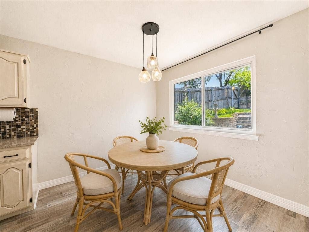 601 Ascot Drive, Euless, TX 76040 Dining area featuring a contemporary three-light pendant fixture, a large window overlooking a fenced backyard, wood-finish flooring, and a portion of a kitchen cabinet with a mosaic tile backsplash