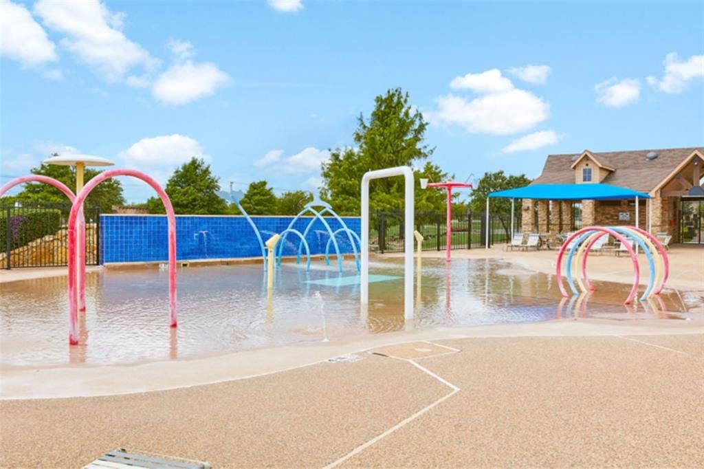 Splash pad featuring multiple water spray elements, a patterned soft-surface ground, and a stone-clad building with a covered patio