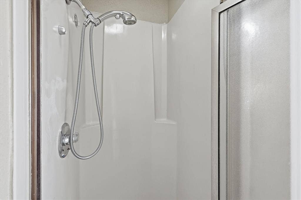 6505 Hughes Drive, Watauga, TX 76148 White shower stall with an integrated bench, featuring a chrome showerhead with a handheld attachment and a textured glass enclosure