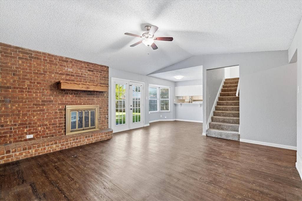 6505 Hughes Drive, Watauga, TX 76148 Spacious living area featuring wood-finish flooring, a brick fireplace with a wood mantle, and a vaulted ceiling with a ceiling fan