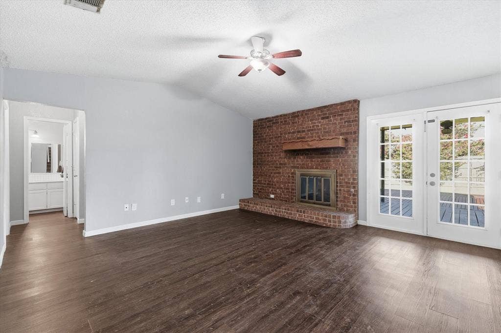 6505 Hughes Drive, Watauga, TX 76148 Spacious living area featuring a brick fireplace, wood-finish flooring, vaulted ceiling, and double French doors opening to a deck