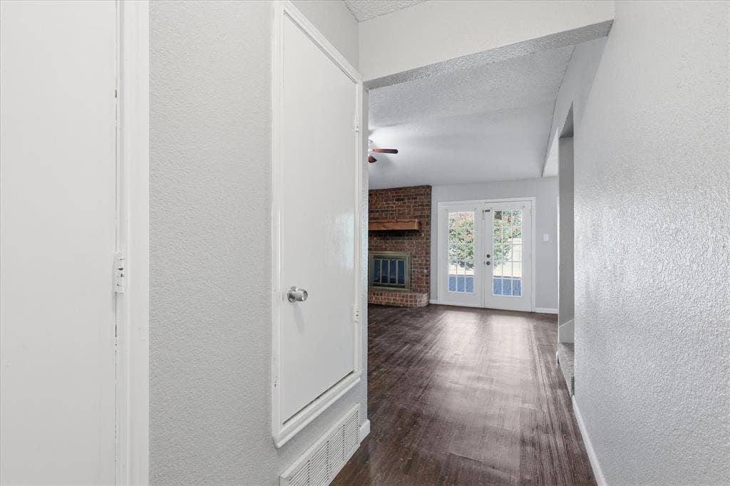 6505 Hughes Drive, Watauga, TX 76148 Hallway with wood-finish flooring leading into a living area featuring a brick fireplace with a wood mantle, double French doors, and a ceiling fan