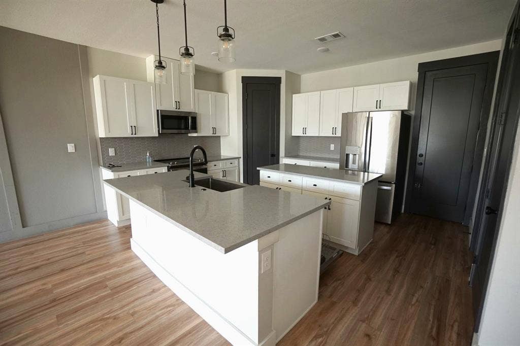 1764 Palladio Loop, Princeton, TX 75407 White cabinetry with matte black hardware, stainless steel appliances, wood-finish flooring, island with integrated sink, and pendant lighting