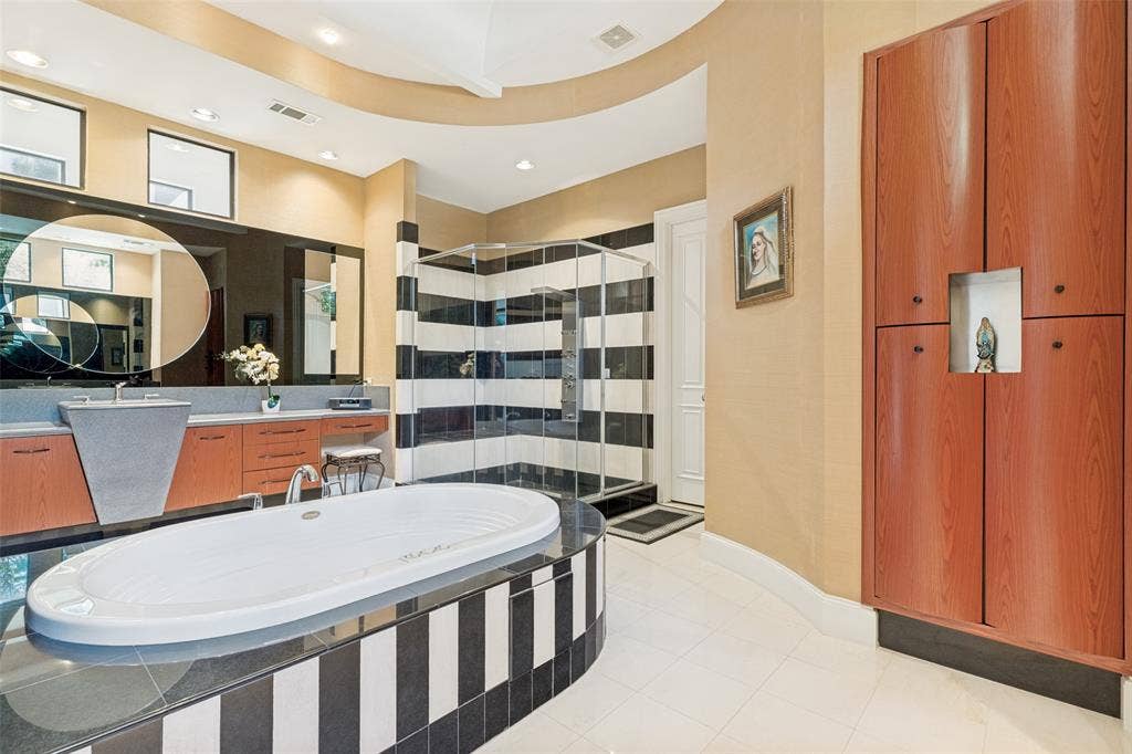 5920 Mcfarland Drive, Plano, TX 75093 Spacious bathroom featuring a white soaking tub with a black and white striped base, a glass-enclosed shower with matching black and white striped tiling, a dual vanity with a unique gray basin and wood-finish cabinetry, and built-in wood-finish storage