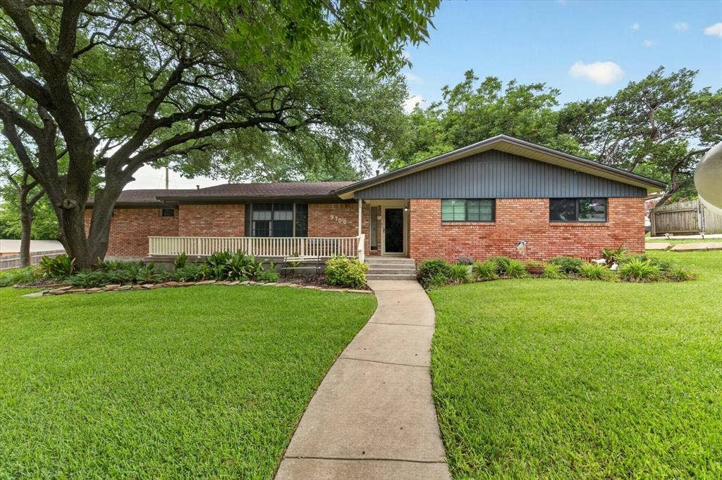 9700 Brookwood Circle, Woodway, TX 76712 Brick exterior home featuring a gabled roofline, a front porch with white railing, a concrete walkway, mature trees, and manicured landscaping