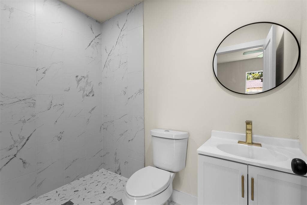 1104 S Brighton Avenue, Dallas, TX 75208 White vanity with gold-tone hardware, a circular mirror, and a walk-in shower with marble-style wall tiles and mosaic floor tiles