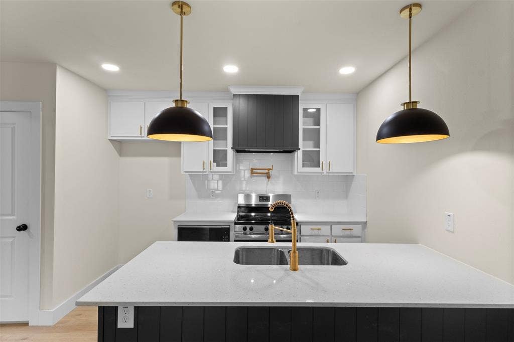 1104 S Brighton Avenue, Dallas, TX 75208 Kitchen island with a light-toned countertop, dual pendant lighting fixtures, a stainless steel sink with a gold-toned faucet, and dark-toned cabinetry