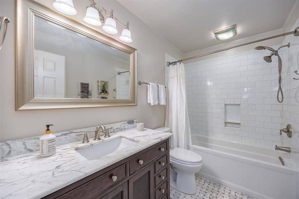 1717 Whitney Drive, Richardson, TX 75082 Bathroom featuring a dark wood-finish vanity with a white rectangular sink, a light-toned countertop, a large framed mirror with overhead lighting, a subway-tiled shower/tub combination with a built-in niche, and a mosaic tile floor
