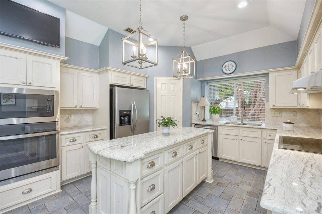 1717 Whitney Drive, Richardson, TX 75082 Kitchen featuring a central island with a light-toned countertop, stainless steel appliances, cream cabinetry with brushed nickel hardware, gray tile flooring, and vaulted ceilings with pendant lighting