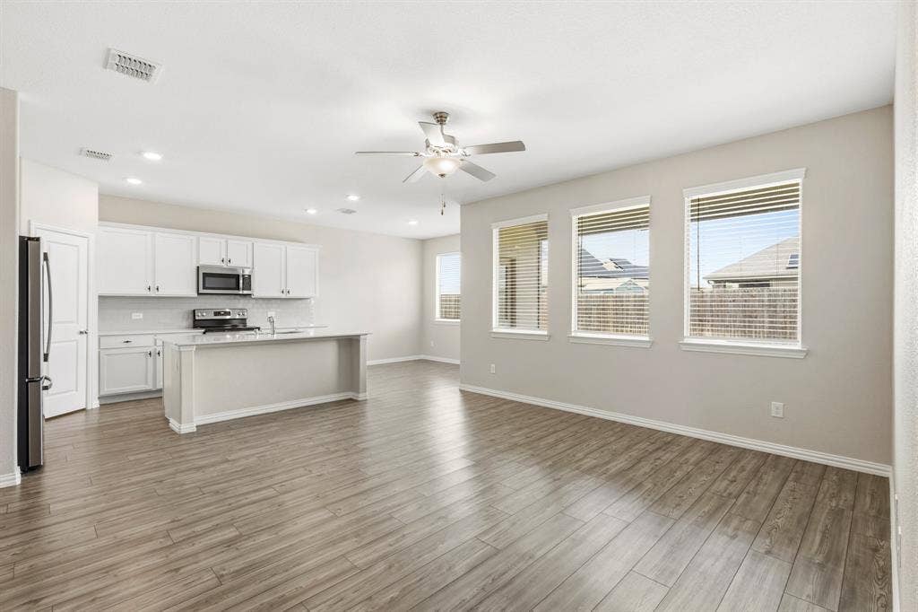 714 Stallings Drive, Princeton, TX 75407 Open-concept space featuring wood-finish flooring, a kitchen island with a built-in sink, white cabinetry with stainless steel appliances, and multiple windows with blinds