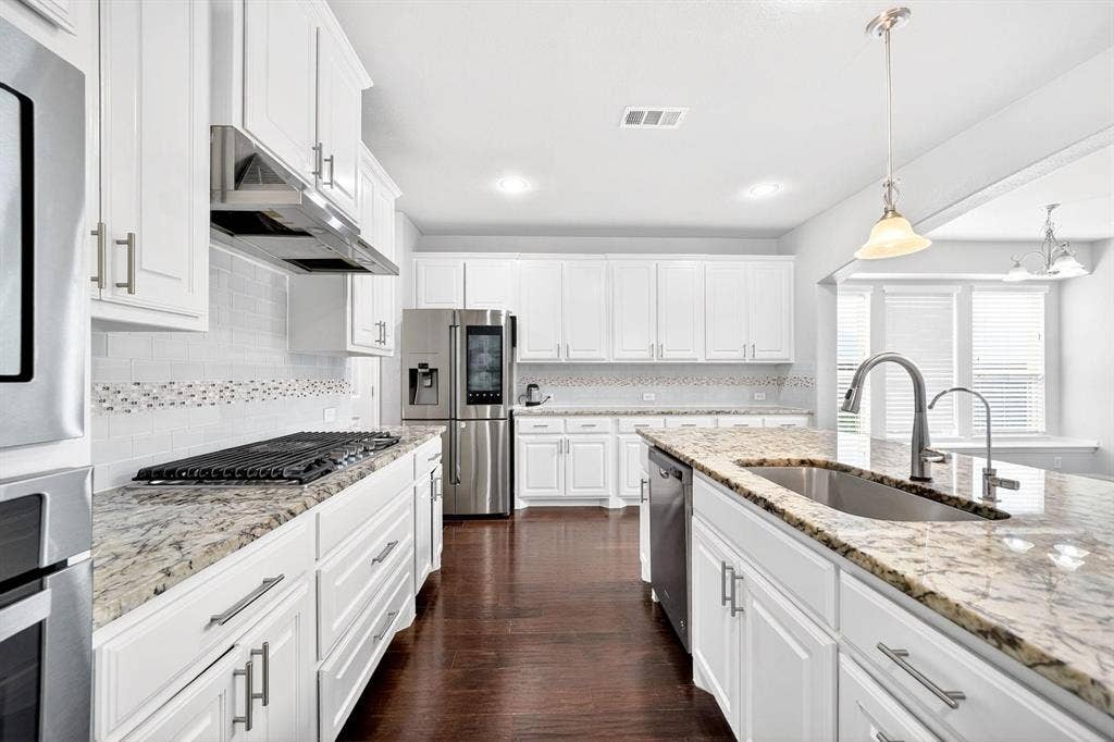 Gourmet kitchen featuring white cabinetry, light granite countertops, stainless steel appliances, a gas range with an overhead vent hood, and a large center island with an undermount sink