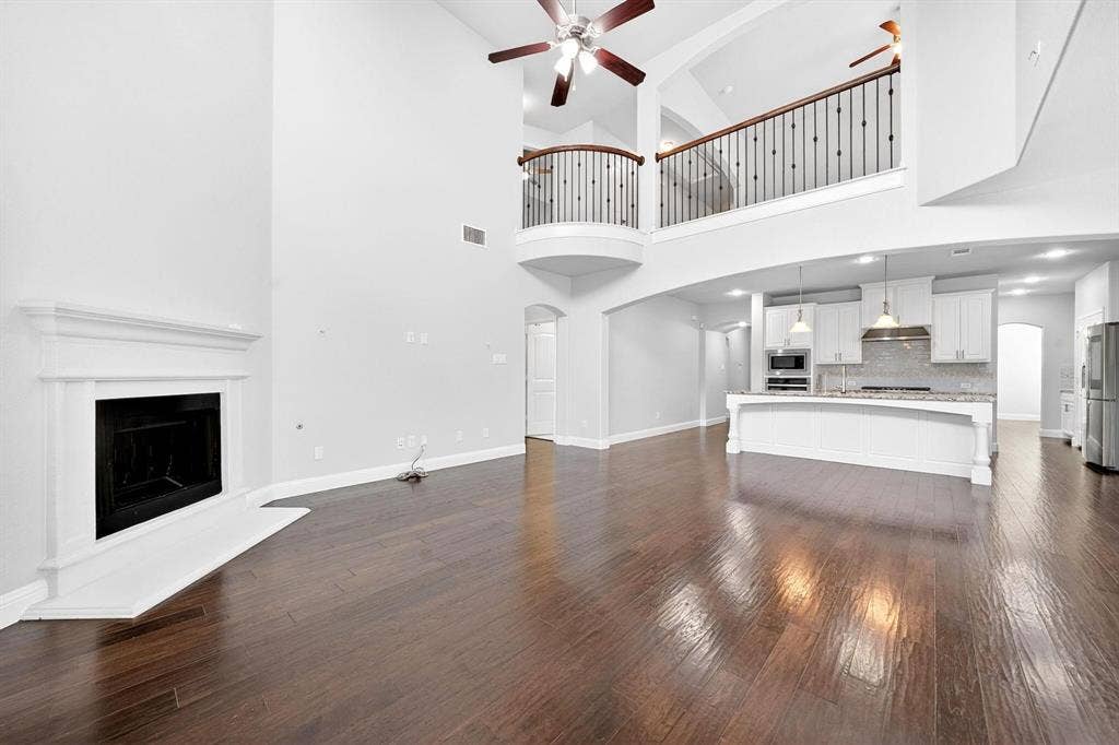 Open-concept living space featuring high ceilings, wood-finish flooring, a white fireplace with mantle, a second-story balcony with wrought iron spindles, and a kitchen island with pendant lighting
