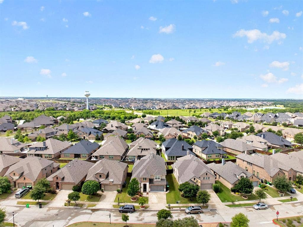 Sprawling residential community with a mix of brick and stone-clad homes, diverse rooflines, and mature landscaping, all set against a distant water tower and extensive green spaces