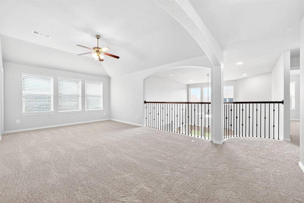 Spacious upper-level area featuring light-toned carpeting, three windows with blinds, a ceiling fan with wood-finish blades, and an open railing with dark balusters