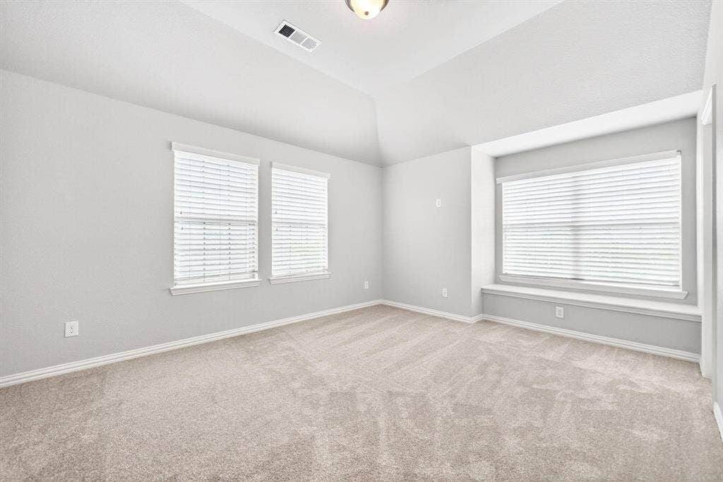 Spacious room featuring vaulted ceilings, light-toned carpeting, multiple windows with blinds, a built-in window seat, and neutral wall paint