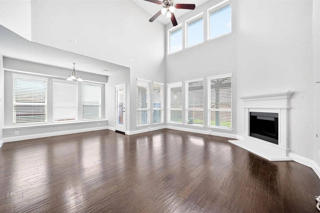 Spacious living area featuring high ceilings, wood-finish flooring, a fireplace with a white mantel, multiple windows with blinds, and a ceiling fan