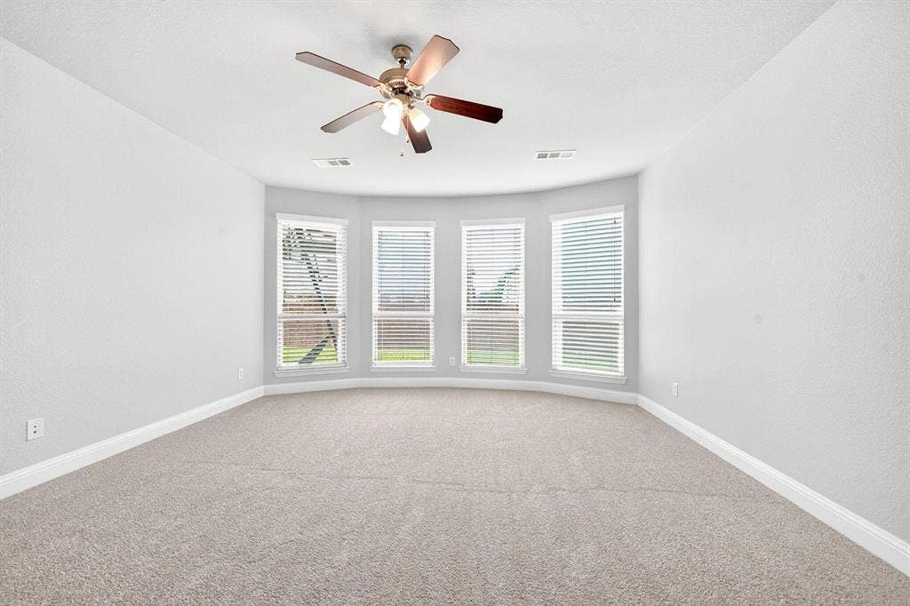 Room featuring a bay window with four individual windows, plush carpeting, light gray walls, white baseboards, and a ceiling fan with integrated lighting