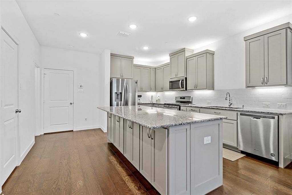 616 Collier Drive, Plano, TX 75075 Spacious kitchen featuring an extended island with granite countertops, stainless steel appliances, light gray shaker cabinetry, a white subway tile backsplash, and dark wood-finish flooring