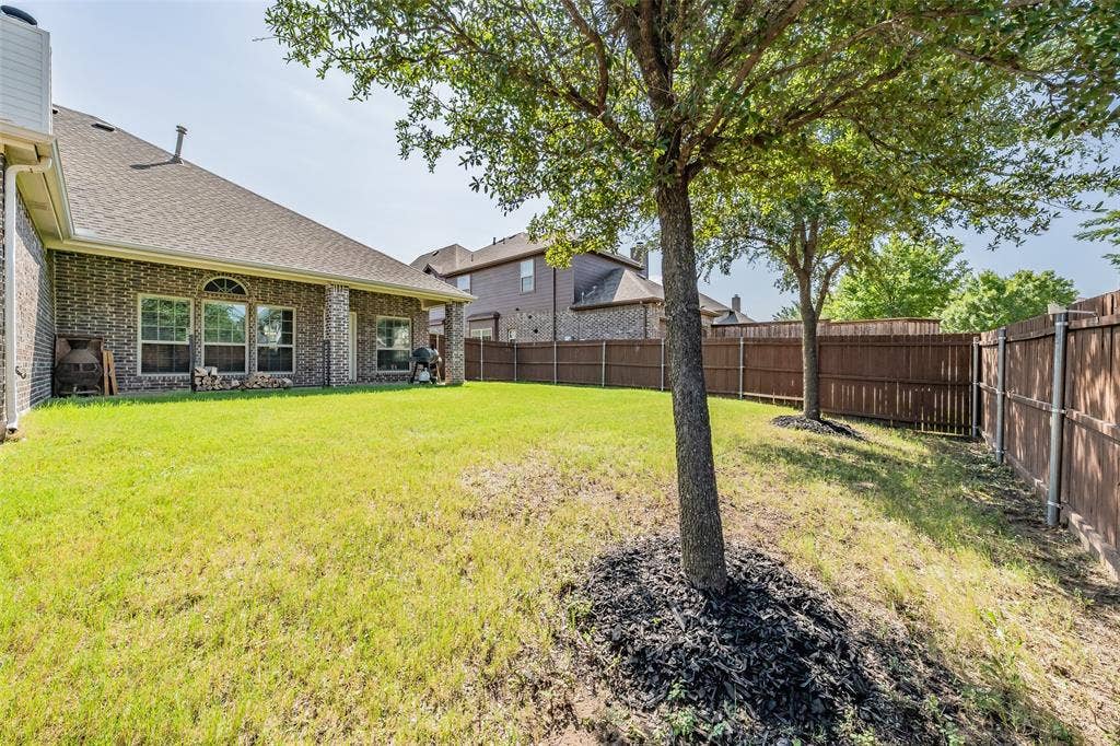 Spacious backyard featuring a brick exterior, covered patio, multiple mature trees, and privacy fencing