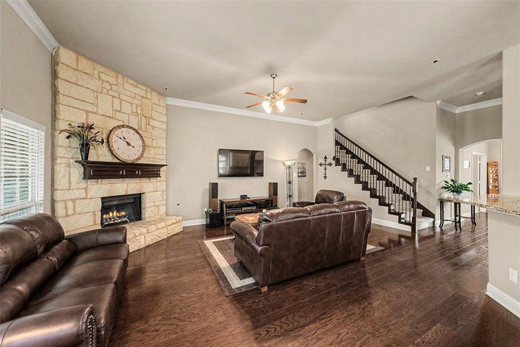 Spacious living area featuring a floor-to-ceiling stacked stone fireplace with a wood mantel, rich wood-finish flooring, a contemporary ceiling fan, and a prominent staircase with dark wood handrails and white balusters