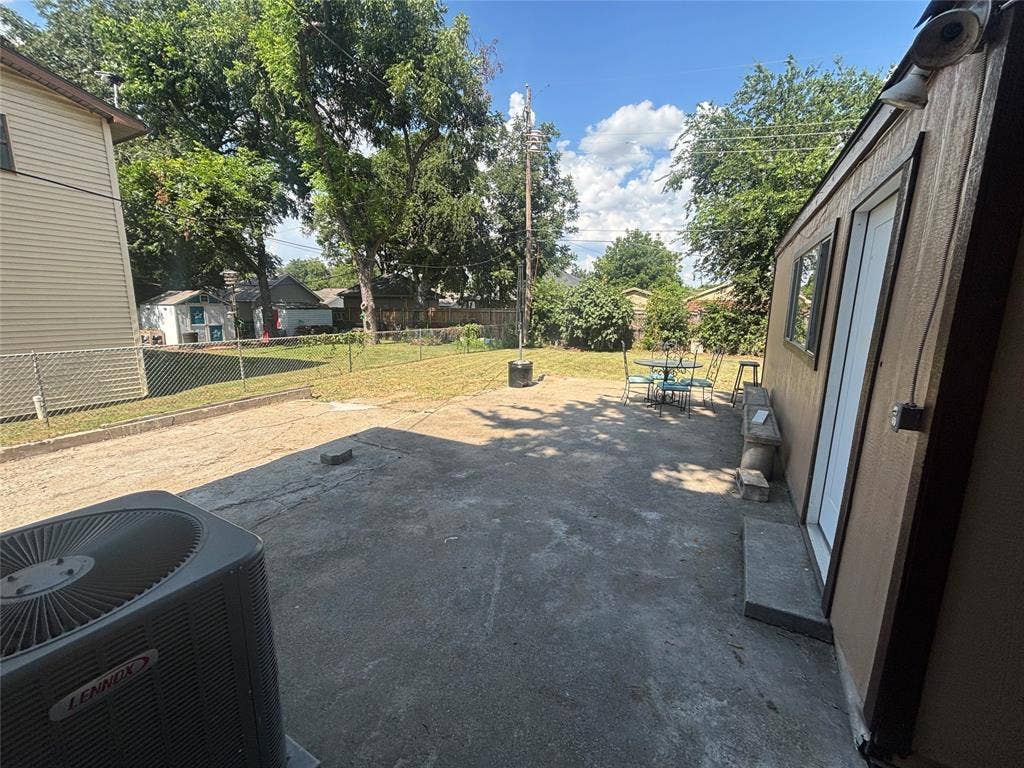 910 NE 31st, Grand Prairie, TX 75050 Spacious concrete patio area featuring a Lennox HVAC unit, a separate exterior structure with a window and door, and a chain-link fence bordering a grassy area with mature trees