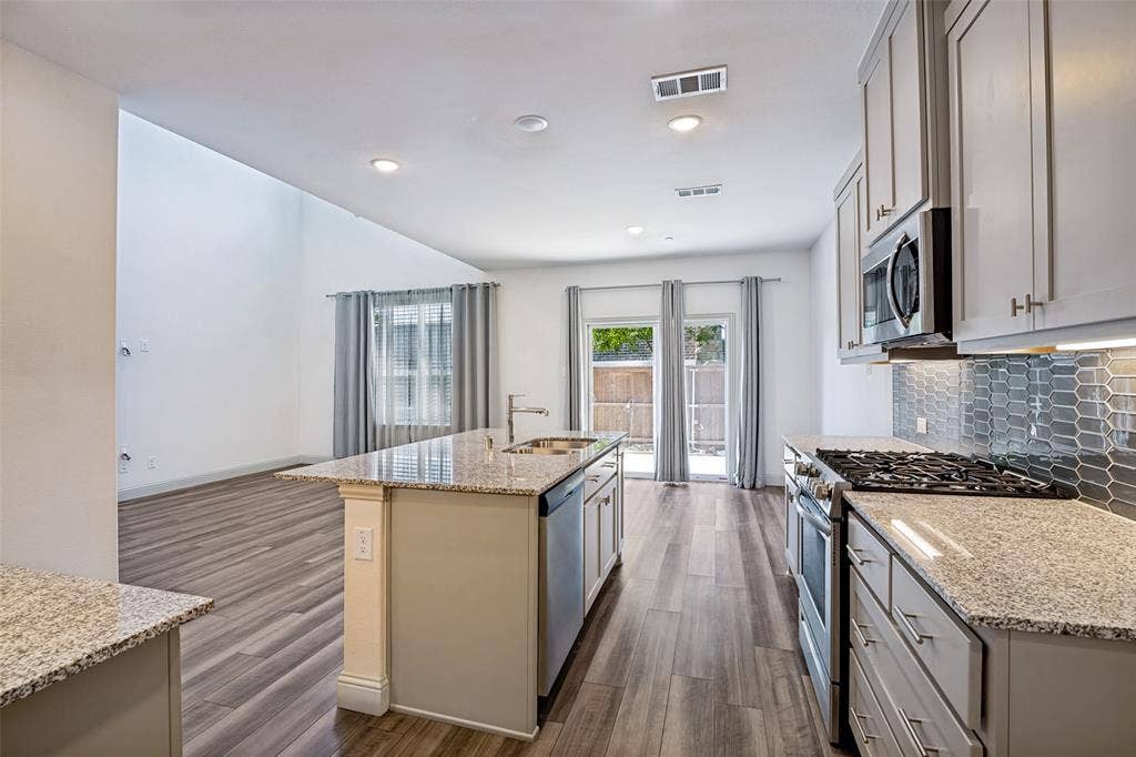 1006 Emil Place, Allen, TX 75013 Kitchen with wood-finish flooring featuring granite countertops, stainless steel appliances, a tile backsplash, and an island with a built-in sink