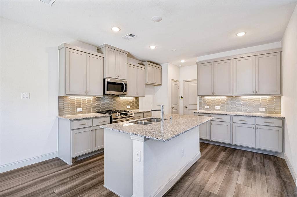 1006 Emil Place, Allen, TX 75013 Open-concept kitchen featuring an island with a double sink, light grey cabinetry, stainless steel appliances, granite countertops, and wood-finish flooring