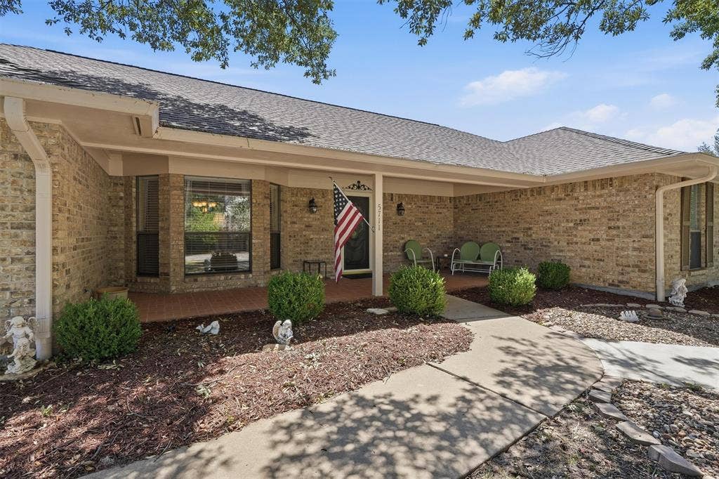5711 Cortez Drive, Granbury, TX 76049 Brick exterior with a covered porch, featuring a bay window, established landscaping, a concrete walkway, and a gray shingle roof