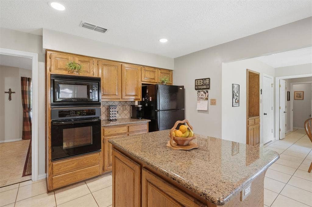 5711 Cortez Drive, Granbury, TX 76049 Kitchen featuring a granite-topped island, wood cabinetry, tile flooring, and black built-in appliances