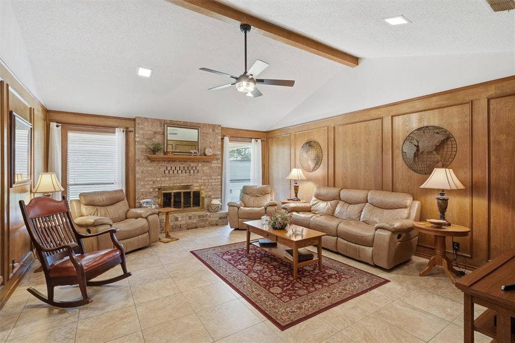 5711 Cortez Drive, Granbury, TX 76049 Spacious living area featuring a vaulted ceiling with exposed wood beams, a brick fireplace, light-toned tile flooring, and wood paneling on the walls