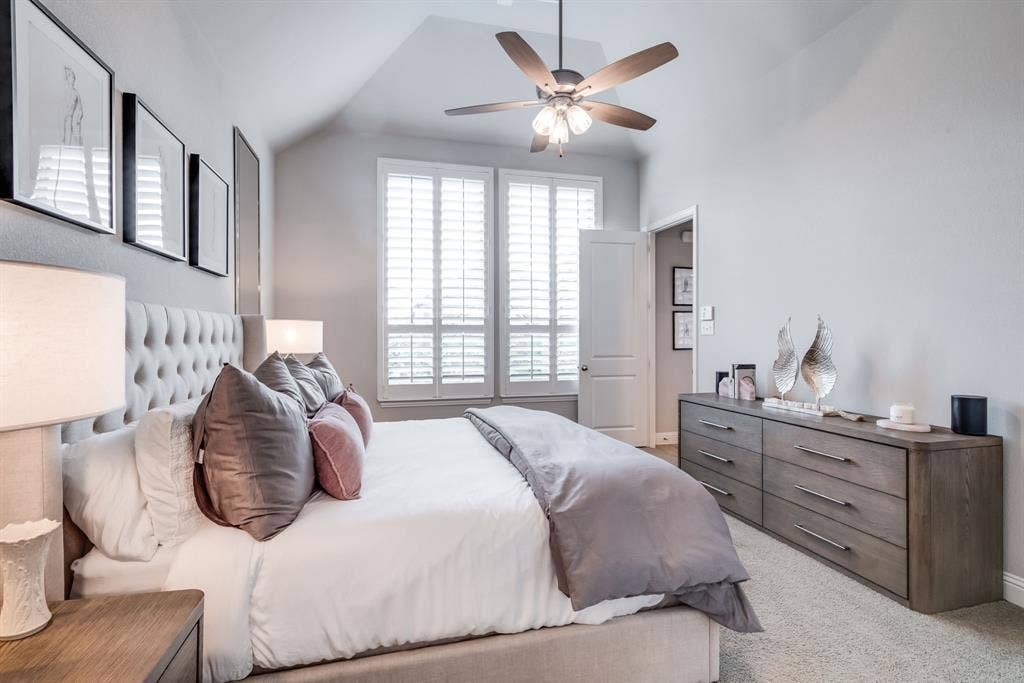Vaulted ceiling room featuring a tufted upholstered headboard, dual windows with plantation shutters, ceiling fan, and carpeted flooring