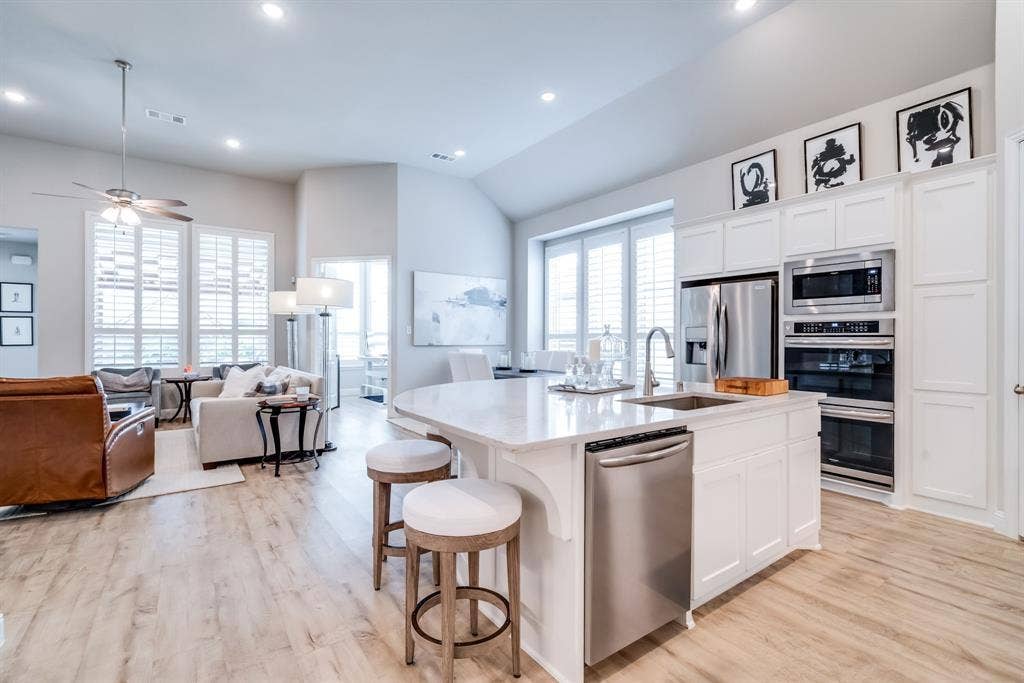 Open-concept living space featuring a kitchen island with seating, stainless steel appliances, wood-finish flooring, vaulted ceilings, and extensive window shutters