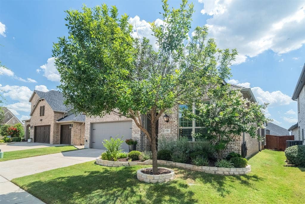 Brick exterior home featuring a two-car garage, established front yard landscaping with stone edging, and mature trees