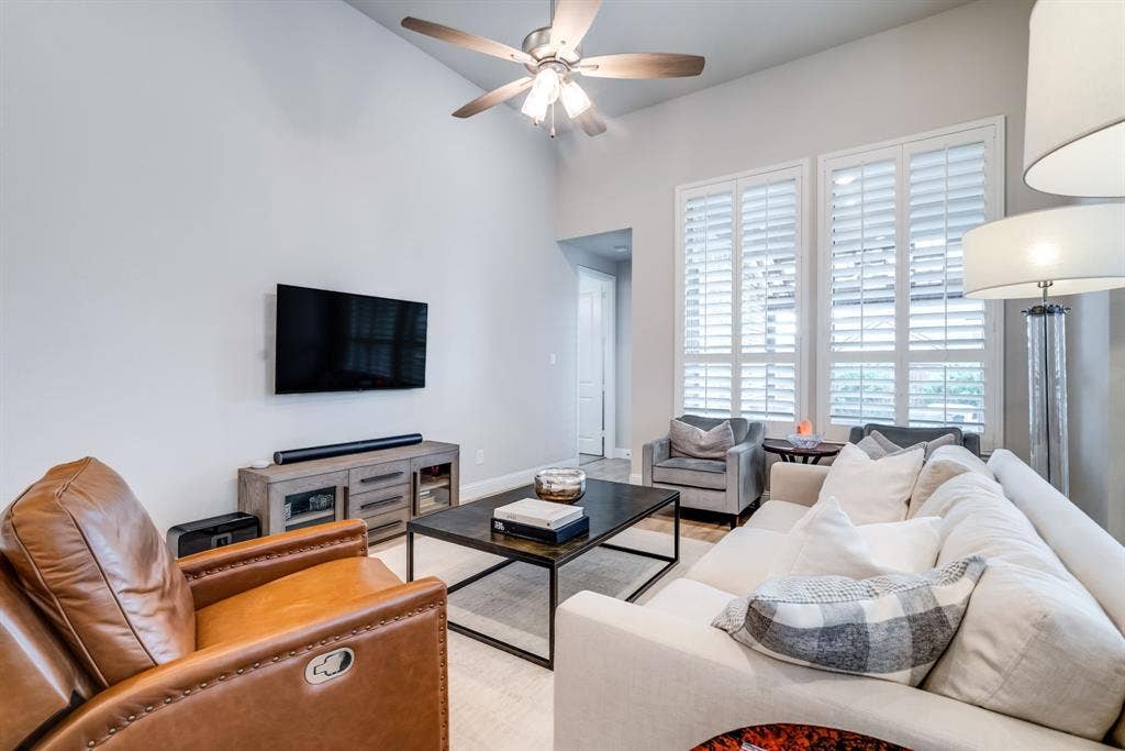 Vaulted ceiling living area featuring twin tall windows with plantation shutters, a ceiling fan with integrated lighting, and light wood-finish flooring