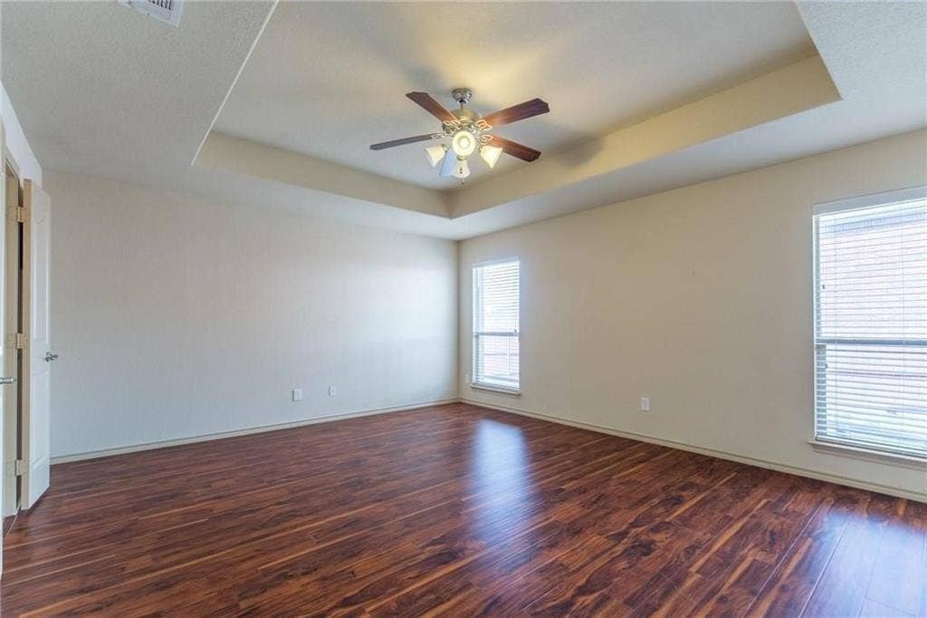 Spacious room featuring wood-finish flooring, a tray ceiling with a ceiling fan and light fixture, and multiple windows with blinds
